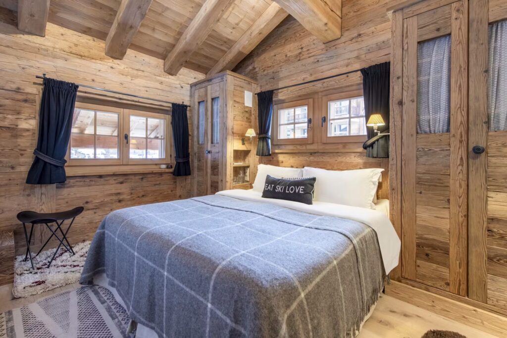 Cozy rustic bedroom with wood walls and ceiling, a large bed with gray plaid blanket, two bedside lamps, small windows with dark curtains, wooden wardrobes, and a decorative pillow reading EAT SKI LOVE. David Pearson Travel