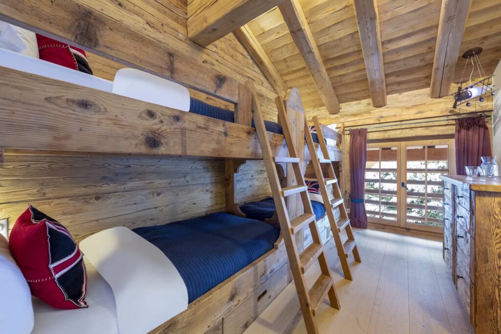 A cozy wooden cabin bedroom with two sets of bunk beds, navy blue bedding, and decorative pillows. A ladder leads to the top bunks. Sunlight streams in through large windows with red curtains, illuminating the wooden walls and ceiling. David Pearson Travel