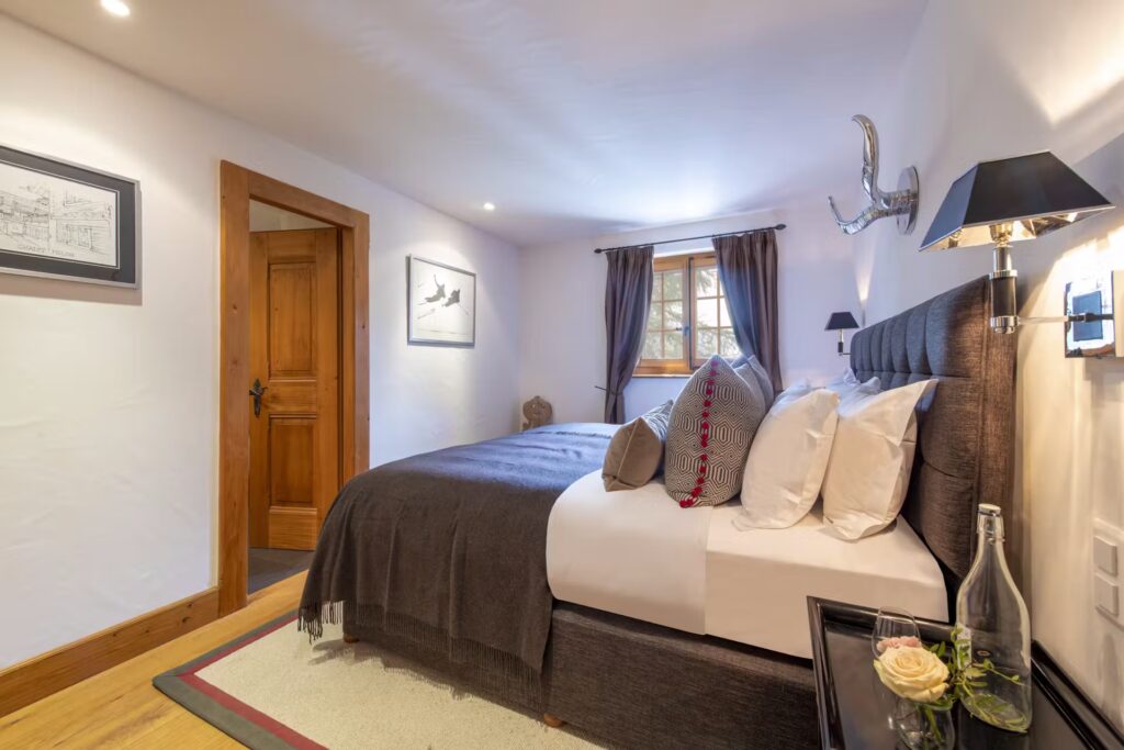 Cozy bedroom with a large bed covered in gray bedding, multiple pillows, bedside tables with lamps, framed artwork on white walls, a wooden door, small window with curtains, and a glass bottle with two glasses on a nightstand. David Pearson Travel