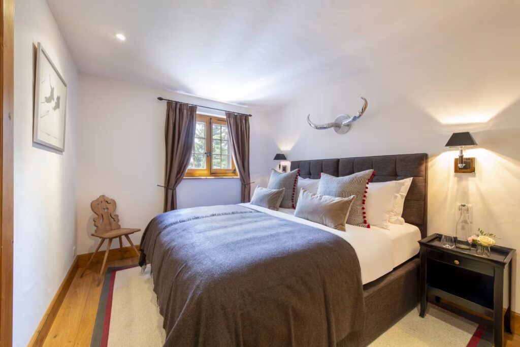 A cozy bedroom with a double bed covered in gray bedding, decorative pillows, a black headboard, wall-mounted lamps, a wooden chair, a window with brown curtains, and a decorative antler wall piece above the bed. David Pearson Travel
