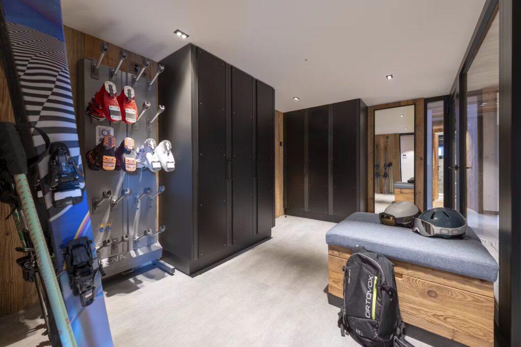 A modern ski locker room with black lockers, a wall-mounted rack holding ski boots and helmets, a padded bench with storage underneath, and a snowboard propped against the wall. The lighting is bright and the decor is sleek. David Pearson Travel