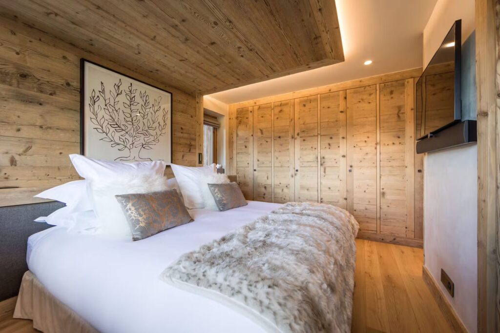 A cozy bedroom with wood-paneled walls and ceiling, featuring a double bed with white linens, decorative pillows, a fur throw, a large wall-mounted TV, and a botanical artwork above the headboard. David Pearson Travel