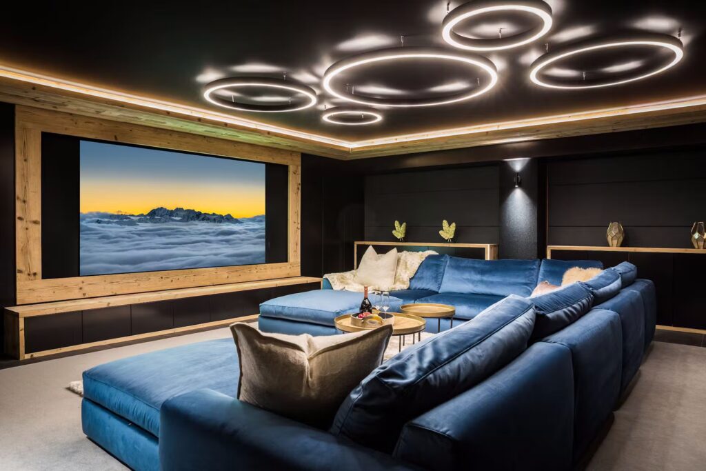 A modern home theater with a large screen showing a mountain scene, surrounded by wooden framing. Blue sectional sofas, round tables, and stylish ceiling lights create a cozy, luxurious atmosphere. David Pearson Travel