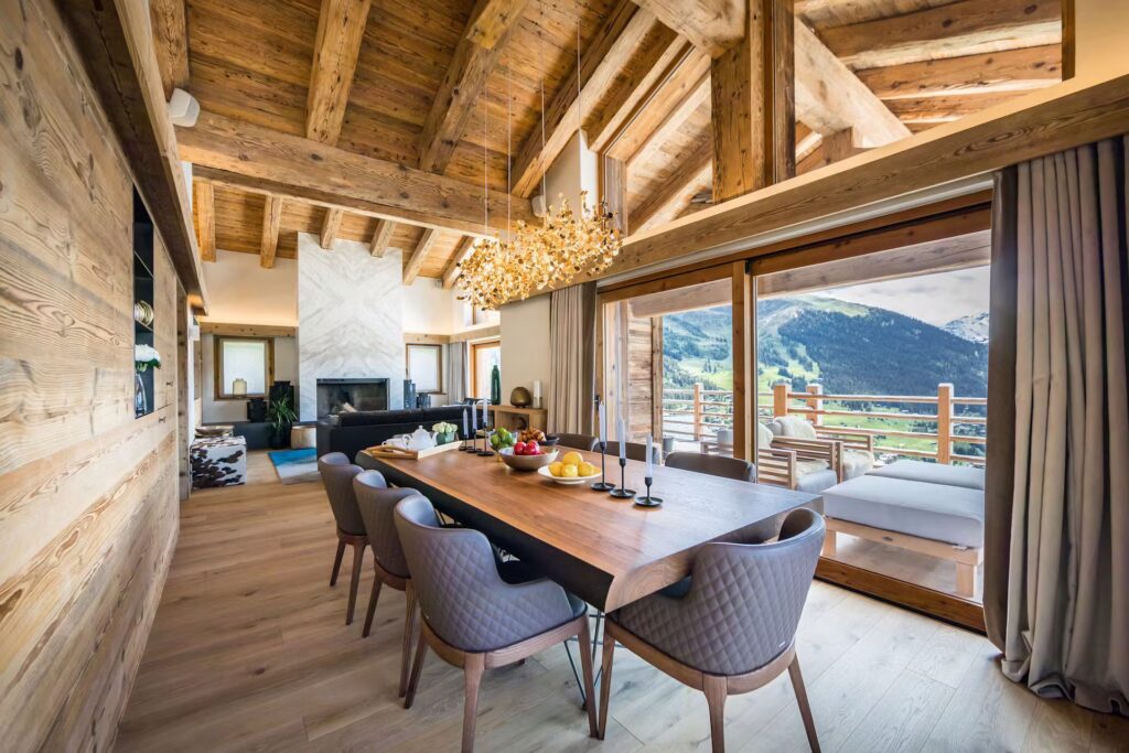 A spacious, modern dining room with a wooden ceiling and walls, large windows showing mountain views, a long dining table with chairs, a chandelier, and a fireplace in the background. David Pearson Travel