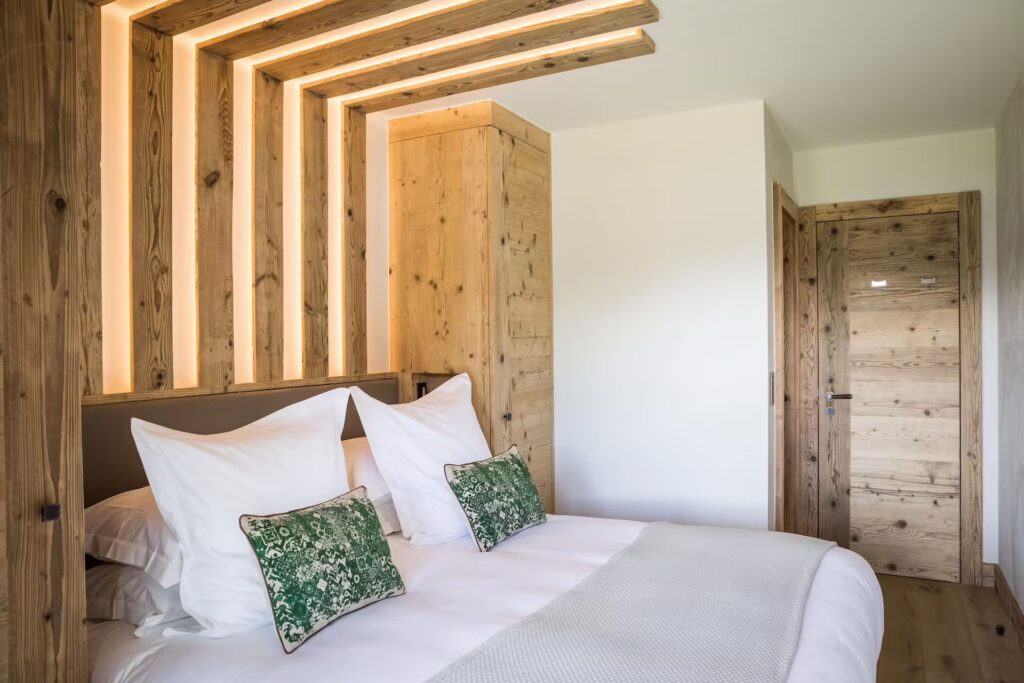 A modern bedroom with wood panel accents, a double bed with white bedding, two green patterned pillows, and white walls. Warm recessed lighting on the ceiling and a wooden door complete the cozy, minimalist decor. David Pearson Travel