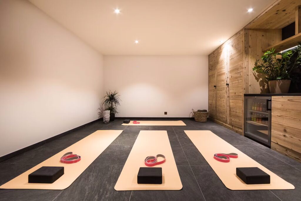 A modern yoga studio with three yellow mats laid out on a dark tile floor, each equipped with a yoga block and strap. Plants, wooden cabinets, and a beverage fridge decorate the minimalist space. David Pearson Travel