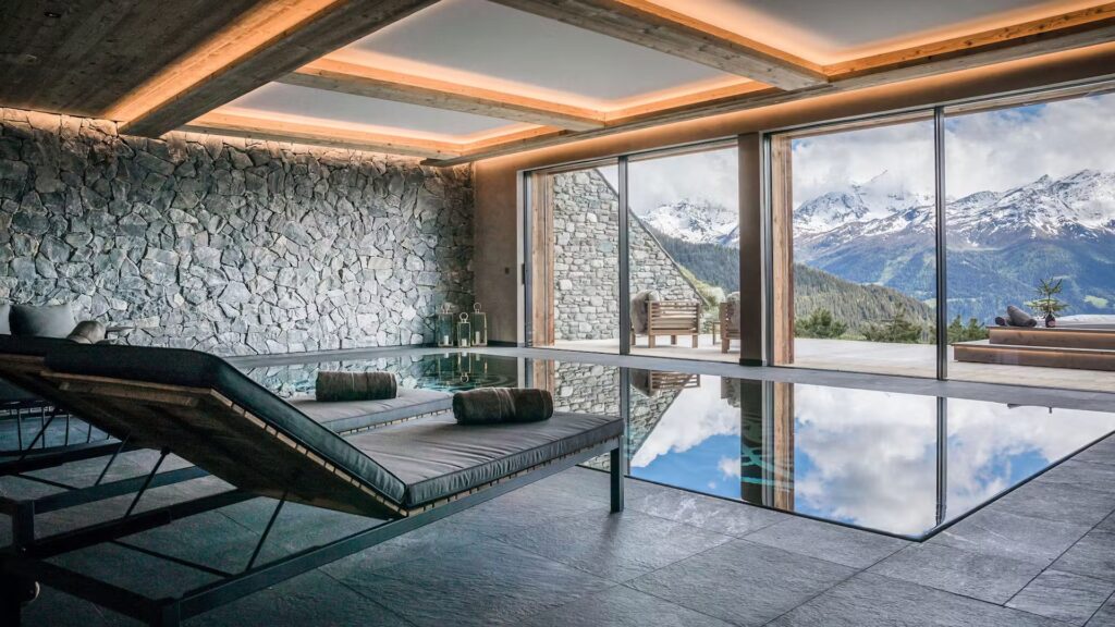 Modern indoor pool room with stone walls, lounge chairs, and floor-to-ceiling windows offering a stunning view of snow-capped mountains and a cloudy sky. The pool reflects the mountains and sky. David Pearson Travel