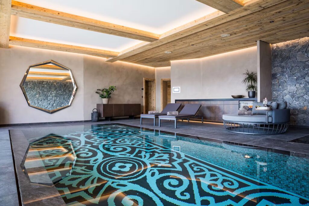 A modern indoor pool with an ornate blue and black mosaic design, surrounded by lounge chairs, a round sofa, stone walls, and a large geometric mirror. The ceiling features wooden beams and recessed lighting. David Pearson Travel