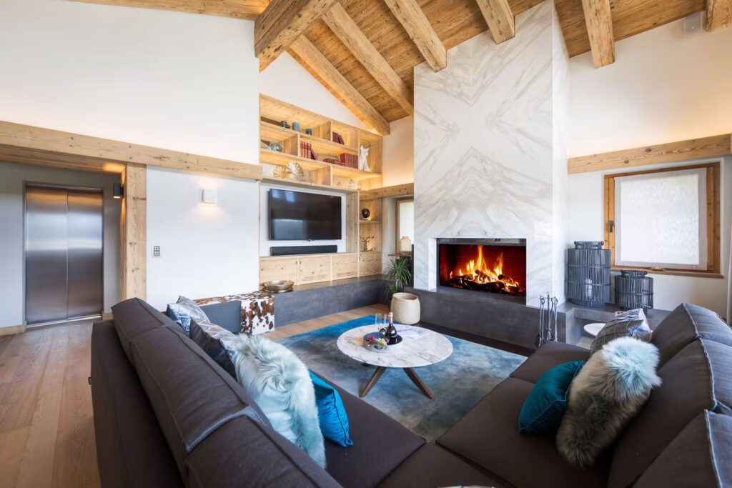 Modern living room with wooden beams, a marble fireplace, cozy gray sectional sofas with blue pillows, a round coffee table, built-in shelves, and a flat-screen TV; elevator and window in the background. David Pearson Travel
