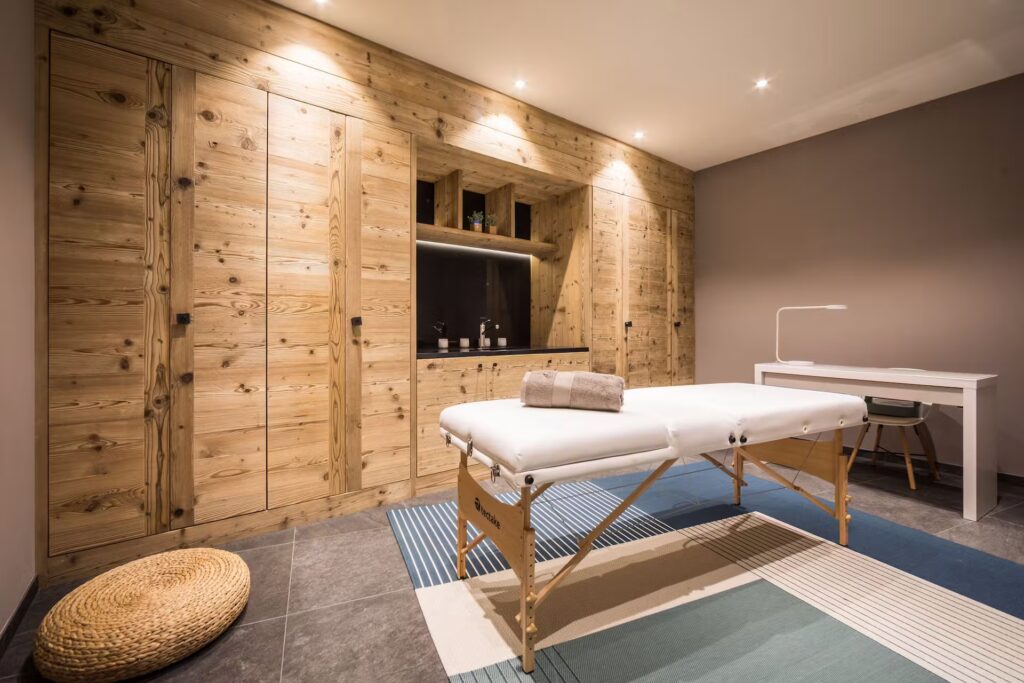 A modern massage room with a wooden wall and built-in shelves, a massage table with towels, a white desk with a lamp, and a woven mat on the floor, all softly lit by ceiling lights. David Pearson Travel