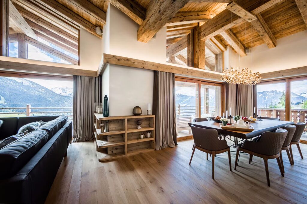 A modern chalet living and dining area with wooden beams, large windows offering mountain views, a black sofa, wooden shelves, dining table with chairs, and a decorative chandelier. Natural light fills the space. David Pearson Travel