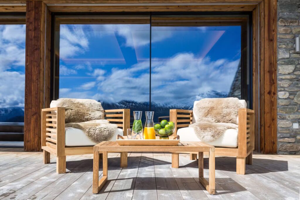 Two wooden chairs with cushions and sheepskin throws sit on a deck facing a glass window reflecting blue sky and clouds. A wooden table holds juice, glasses, and a bowl of green apples. Mountains are visible in the reflection. David Pearson Travel