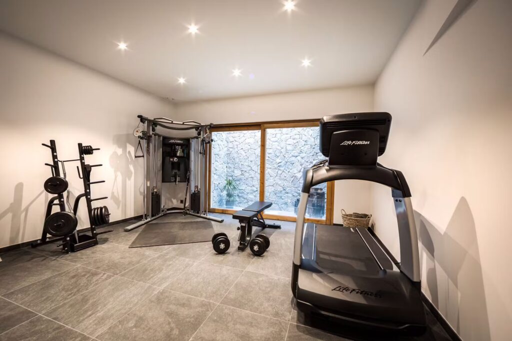 A modern home gym with a treadmill, weight bench, dumbbells, multi-gym machine, rack with weight plates, and large glass doors showcasing a stone wall outside. The room has gray tile flooring and recessed lighting. David Pearson Travel