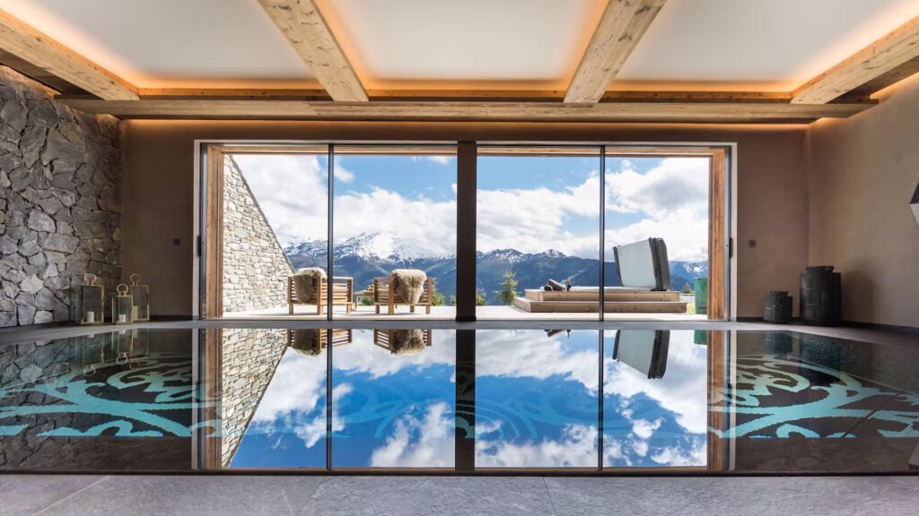 A modern indoor pool with glass doors opening to a patio with lounge chairs, overlooking scenic mountains and blue sky reflected in the water. David Pearson Travel