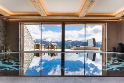 A modern indoor pool with glass doors opening to a patio with lounge chairs, overlooking scenic mountains and blue sky reflected in the water. David Pearson Travel