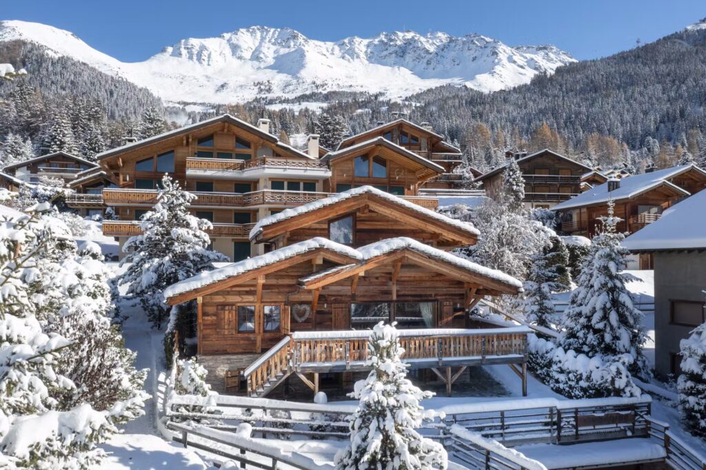 A cluster of wooden chalets surrounded by snow-covered trees sits beneath a backdrop of snowy mountains under a clear blue sky. David Pearson Travel