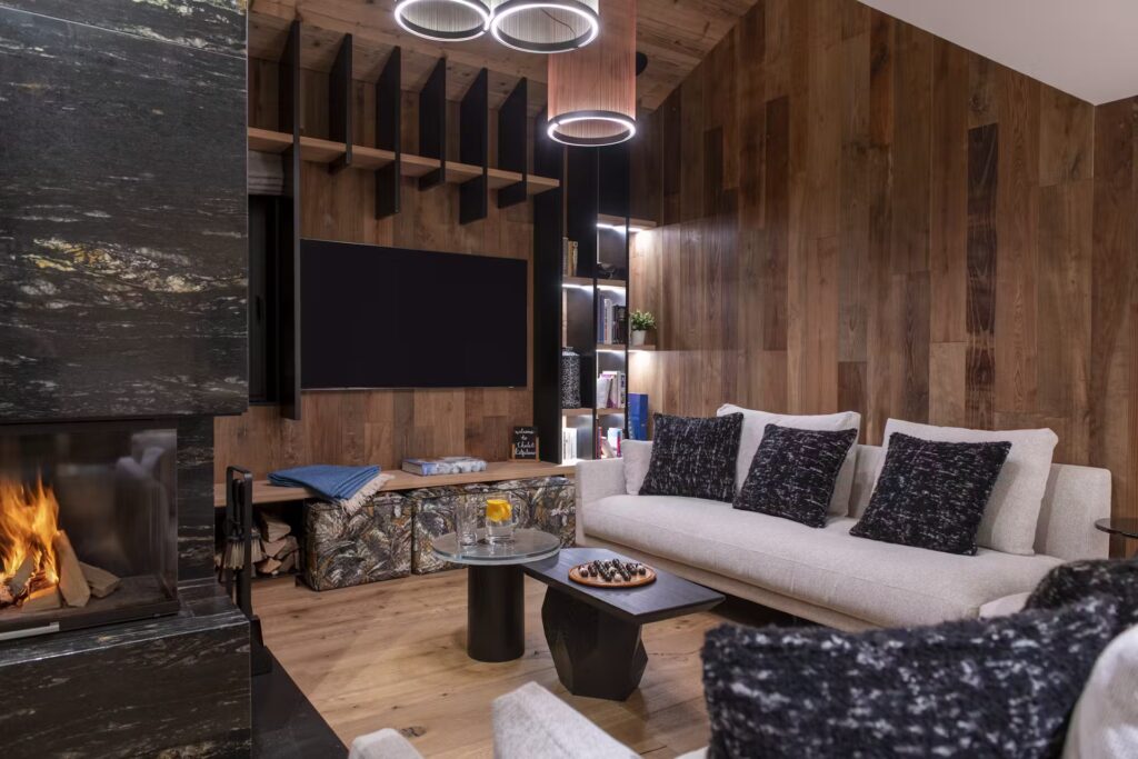 Modern living room with wood-paneled walls, a light gray sofa with dark cushions, a fireplace, a TV, circular ceiling lights, and a black coffee table with snacks and drinks. Shelving and decorative items are visible. David Pearson Travel