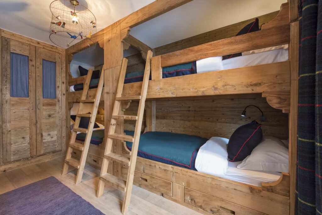 A cozy room with two wooden bunk beds, each with navy bedding and white pillows, a wooden ladder, built-in drawers, a hanging light fixture, and wooden paneled walls. David Pearson Travel