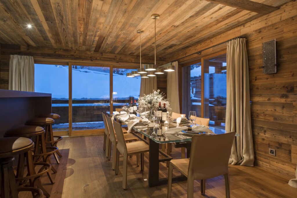A cozy wooden dining room with a glass table set for dinner, beige chairs, hanging lights, and large windows showing a snowy outdoor scene. Neutral curtains frame the windows, and a flower centerpiece decorates the table. David Pearson Travel