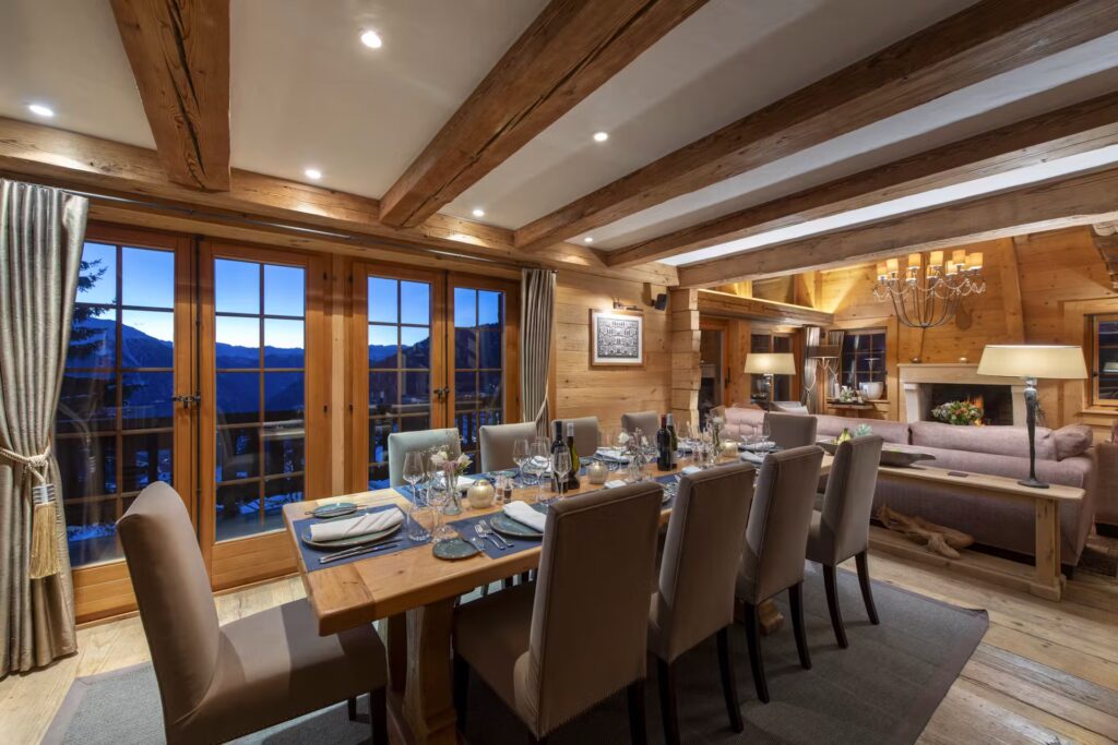 A cozy, elegant dining room with a wooden table set for eight, surrounded by beige chairs. The room features exposed wooden beams, warm lighting, and large windows with mountain views at dusk. David Pearson Travel