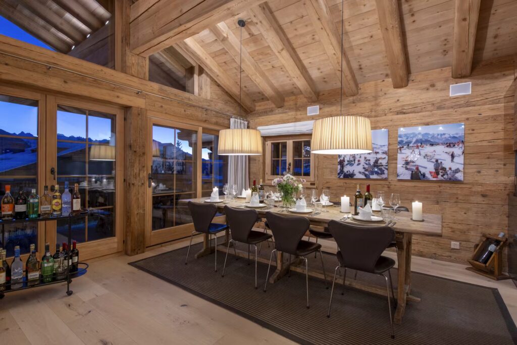 A cozy, modern dining room with wooden walls and ceiling, a long set dining table, candles, two large pendant lights, a bar cart, and large windows showing a twilight mountain view. David Pearson Travel