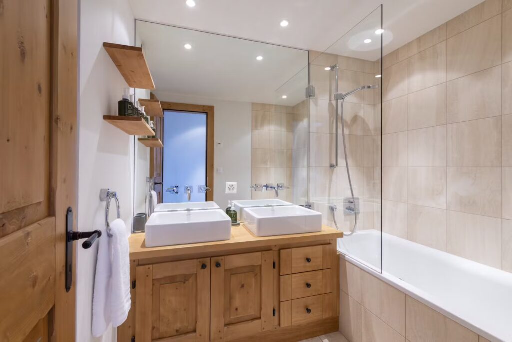 Modern bathroom with light wood cabinets, dual square sinks, a large wall mirror, built-in shelves, and a bathtub with a glass shower screen. Neutral beige tiles cover the walls, and recessed ceiling lights illuminate the space. David Pearson Travel