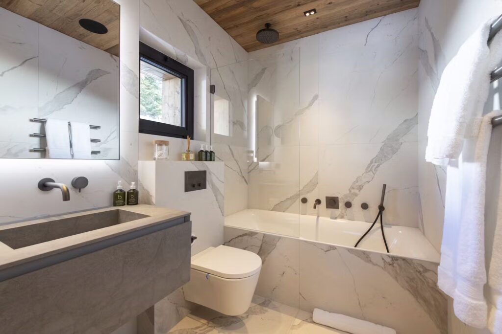 Modern bathroom with marble walls, a gray sink, wall-mounted toilet, bathtub with black fixtures, heated towel rack, large mirror, and a window; wood-paneled ceiling with recessed lights and rain showerhead. David Pearson Travel