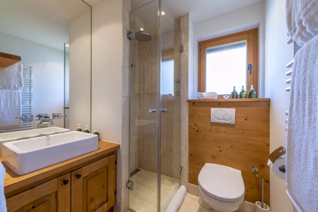 Modern bathroom with a wooden vanity, a square sink, a glass-enclosed shower, white toilet, wall-mounted towel rack, and a window letting in natural light. Bottles and towels are neatly arranged around the room. David Pearson Travel
