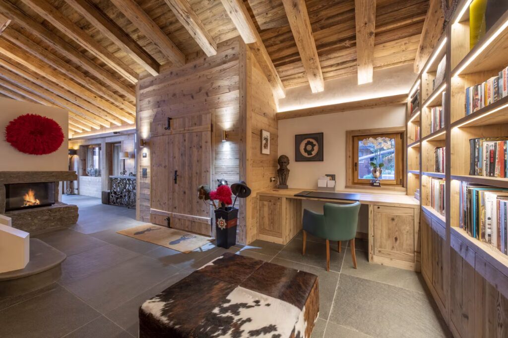 A cozy wooden chalet interior with exposed beams, built-in bookshelves, a desk with a green chair, a cowhide ottoman, a fireplace, and warm ambient lighting. A window shows a view of greenery outside. David Pearson Travel