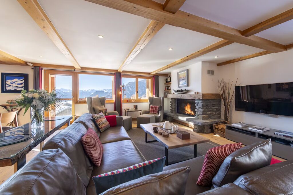 A cozy living room with a large sectional sofa, colorful pillows, a fireplace, a TV, and panoramic windows offering mountain views. Wooden beams and natural light create a warm, inviting atmosphere. David Pearson Travel