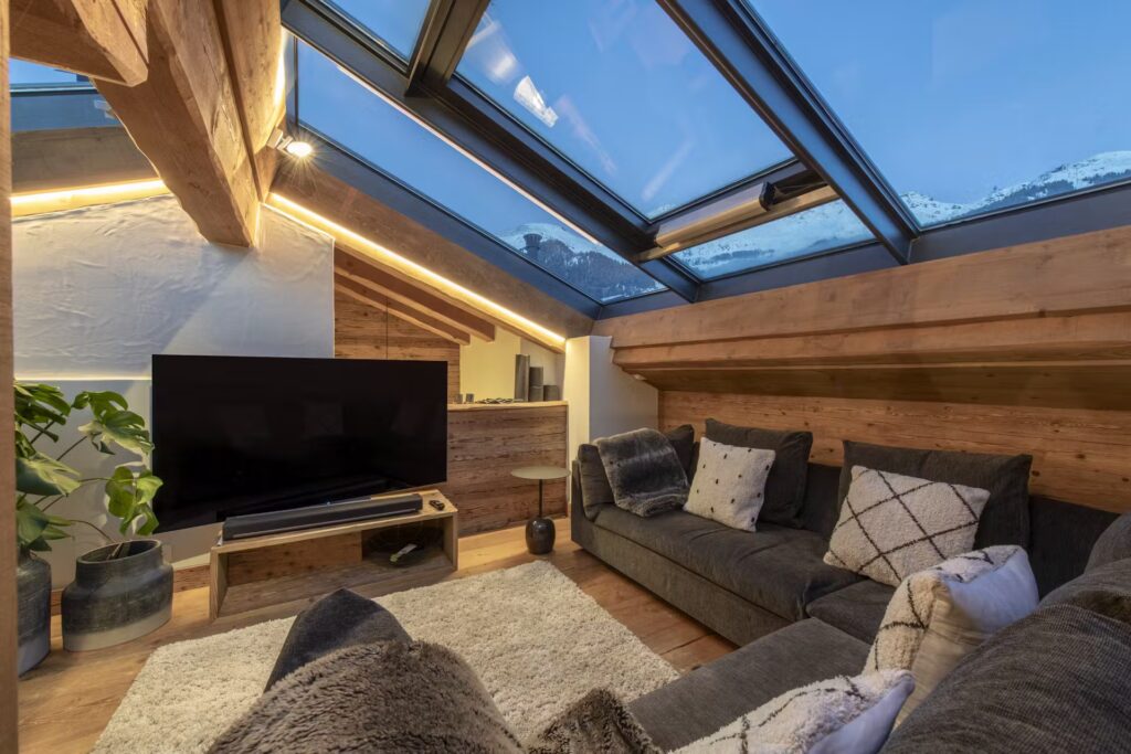 A cozy loft living room with a slanted glass ceiling, wooden beams, a gray sofa with patterned cushions, a large TV, and a view of snowy mountains through the windows at dusk. David Pearson Travel