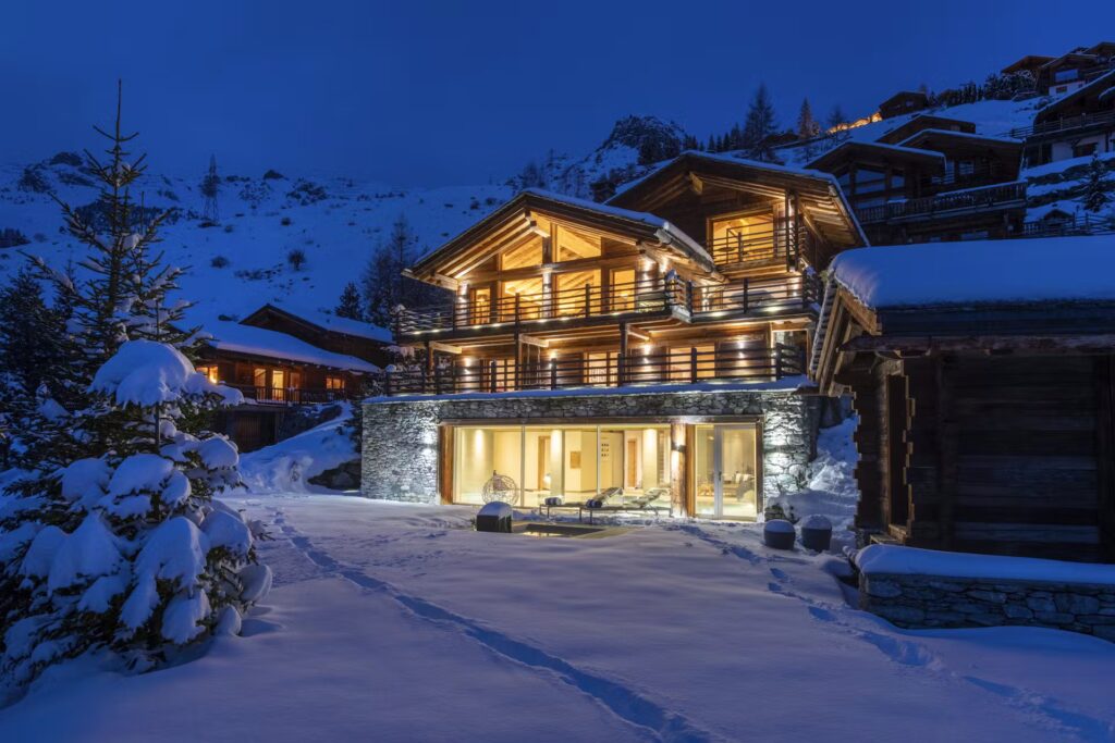 A modern, three-story chalet glows warmly at night, surrounded by snow-covered ground and trees, with mountains and other cabins in the background under a deep blue sky. David Pearson Travel