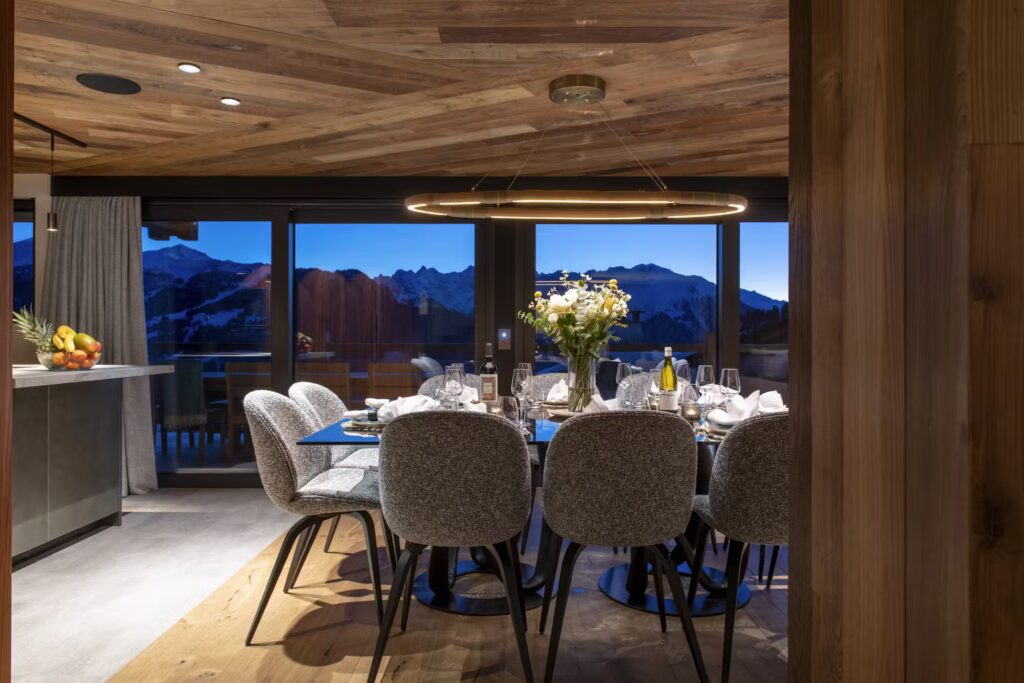 A modern dining room with a wooden ceiling, a large table set for eight, elegant gray chairs, and a circular chandelier. Floor-to-ceiling windows reveal a mountain view at dusk. A kitchen with fruit is visible on the left. David Pearson Travel