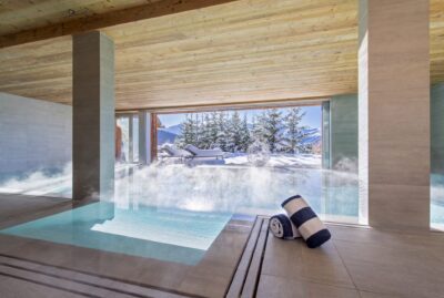 A modern indoor pool with steam rising from the water, next to a large window overlooking snowy mountains and pine trees. Two rolled towels with blue and white stripes rest on the tiled floor. David Pearson Travel