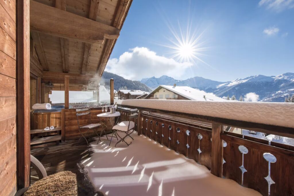 A wooden balcony covered in snow overlooks snowy rooftops and distant mountains under a bright, sunny blue sky. There is a small table with two chairs, and steam rises from a hot tub in the corner. David Pearson Travel