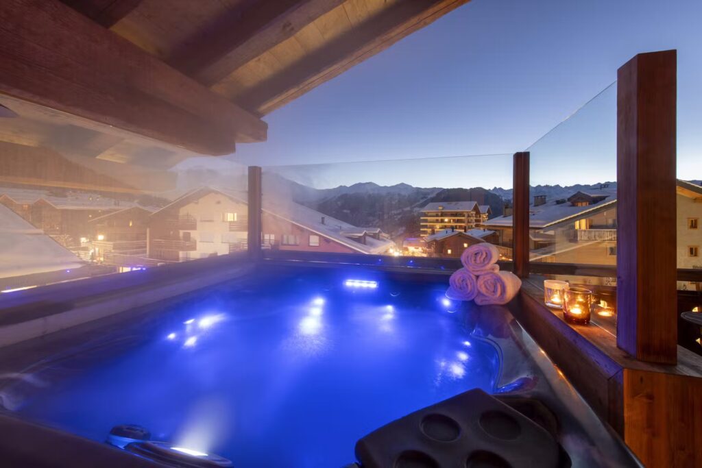 A glowing outdoor hot tub with blue lights sits on a balcony overlooking snowy rooftops and mountains at dusk, with rolled towels and lit candles on the side. David Pearson Travel