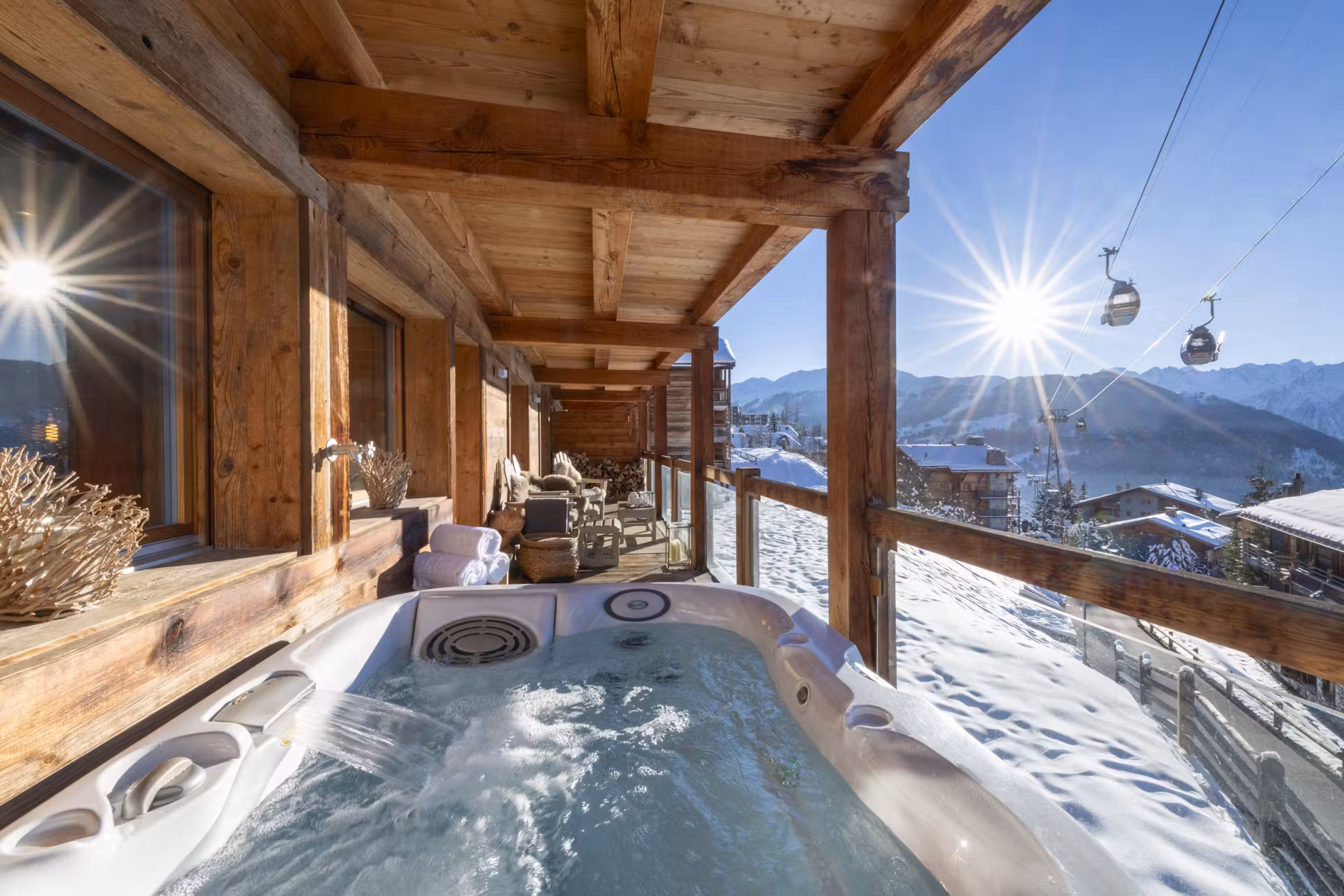 A wooden chalet balcony features a bubbling hot tub overlooking snowy mountains and ski gondolas under a bright blue sky with sunlight streaming in. Cozy seating and rustic decor complete the inviting winter scene. David Pearson Travel