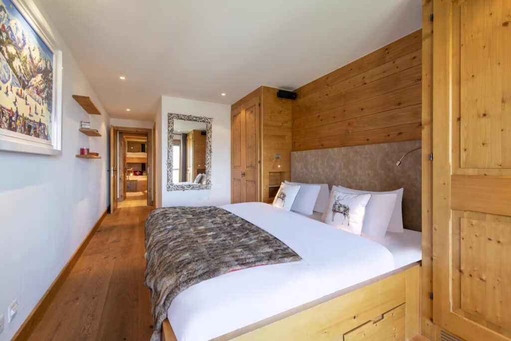 A cozy bedroom with a large bed dressed in white linens and a fur blanket, wooden walls and floors, a mirror, shelves, and a doorway leading to another room. A framed artwork hangs on the white wall. David Pearson Travel