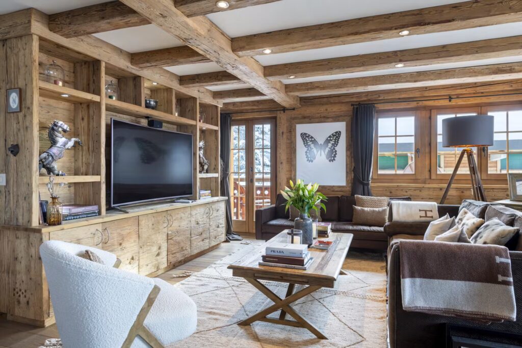 A cozy living room with rustic wooden beams, a large TV, brown sofas, a white chair, a coffee table with books and flowers, built-in shelving, and a large butterfly artwork on the wall near sunny windows. David Pearson Travel