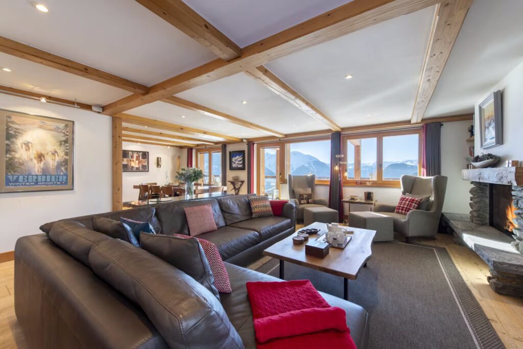 Spacious living room with large leather sofa, armchairs, wooden beams, stone fireplace, and mountain views through wide windows; dining area visible in the background. Cozy, modern, and well-lit. David Pearson Travel