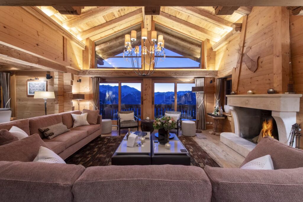 Cozy, spacious living room with wood paneling, plush sofas, a fireplace, large windows showing mountain views at sunset, and a chandelier hanging from a vaulted ceiling. David Pearson Travel