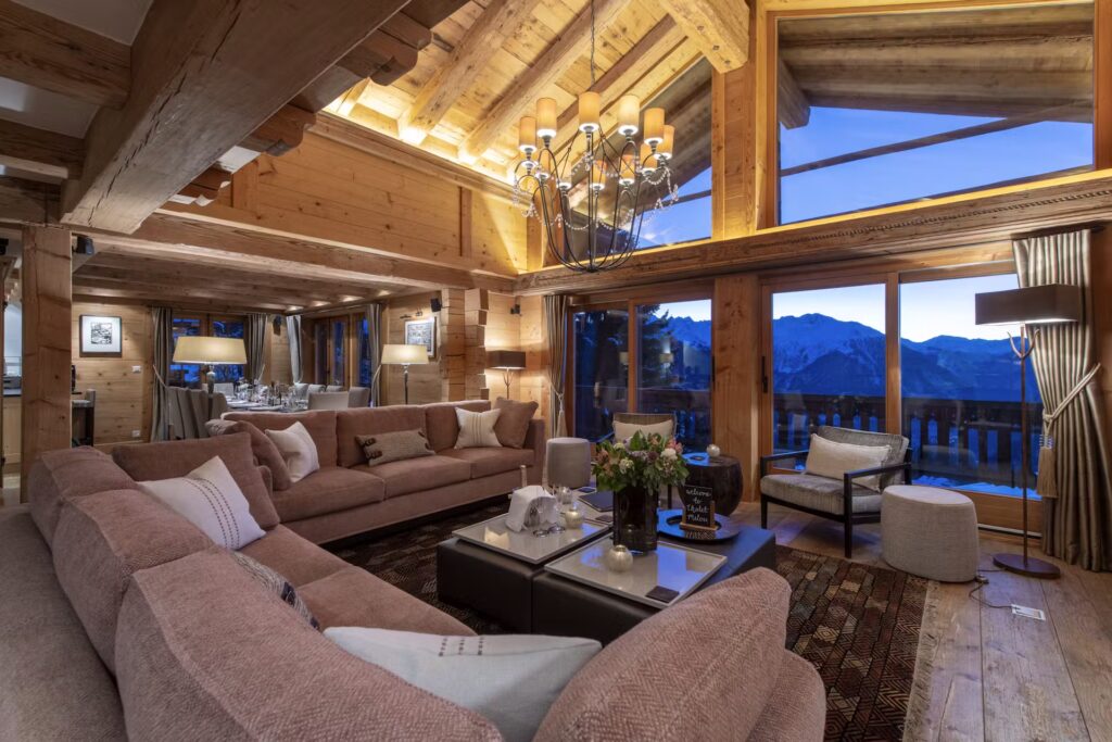 A spacious, cozy living room with wooden beams, large sofas, a chandelier, and floor-to-ceiling windows revealing a mountain view at dusk. A dining area is visible in the background, bathed in warm lighting. David Pearson Travel