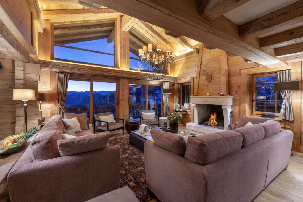 A cozy wooden chalet living room with large windows, mountain views, plush sofas, a fireplace, warm lighting, and a high ceiling with exposed beams and a chandelier. David Pearson Travel