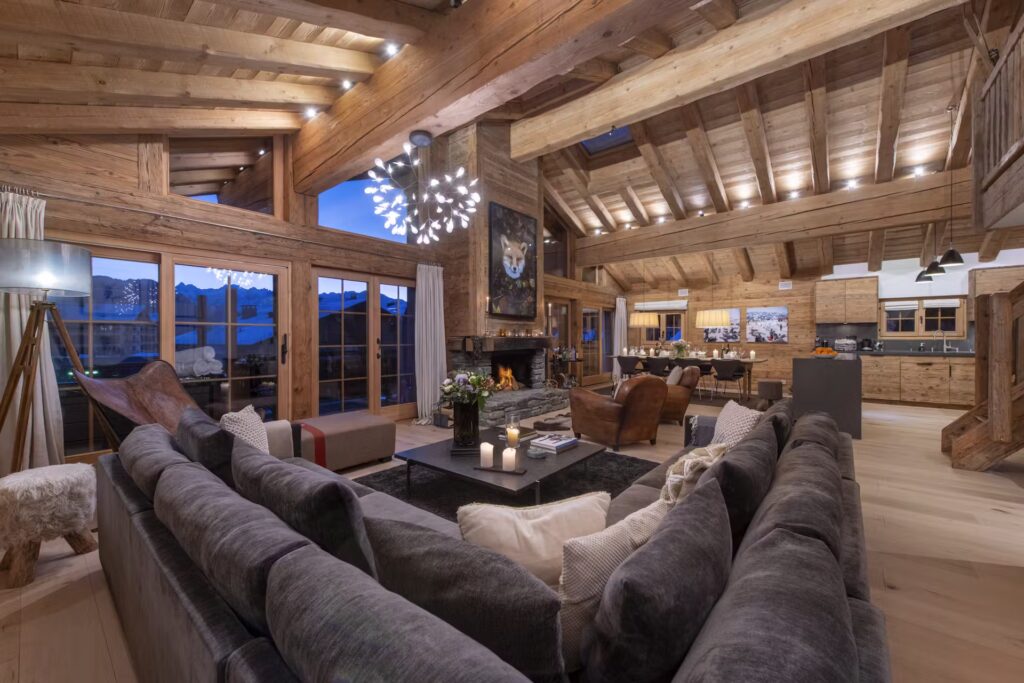 Spacious rustic living room with exposed wooden beams, a cozy gray sectional sofa, modern chandelier, stone fireplace, and large windows showing mountain views at dusk. Open layout leads to dining and kitchen areas. David Pearson Travel