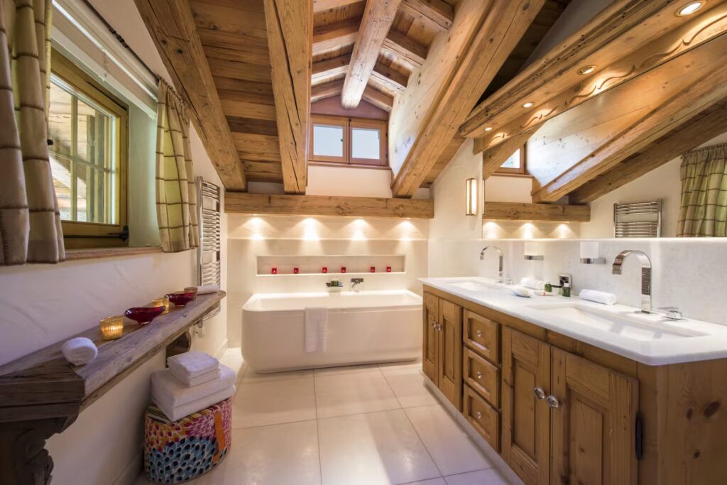 A cozy, rustic bathroom with exposed wooden beams, a large bathtub, dual sinks with wooden cabinets, soft lighting, and decorative accents like candles and neatly folded towels. David Pearson Travel