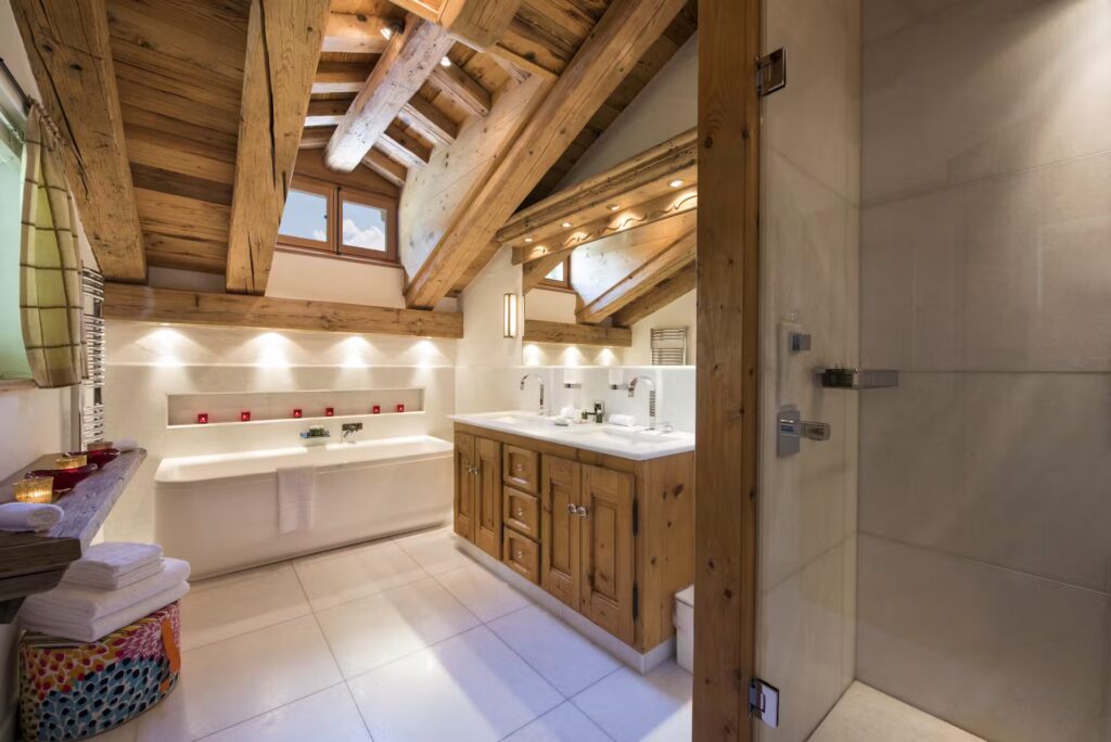 Modern rustic bathroom with exposed wooden beams, a large bathtub, wooden vanity with double sinks, glass shower, white tile floor, and soft ambient lighting. Towels and candles add cozy touches. David Pearson Travel