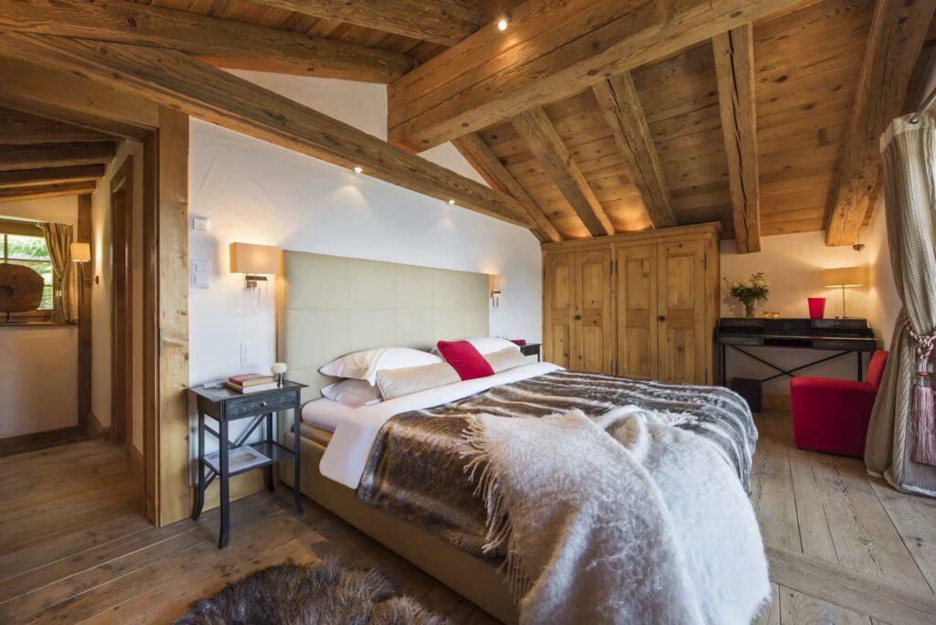 Cozy bedroom with rustic wooden beams, a large bed with fur throw, bedside tables, an armoire, and a desk. Warm lighting and natural wood accents create a welcoming, alpine atmosphere. David Pearson Travel