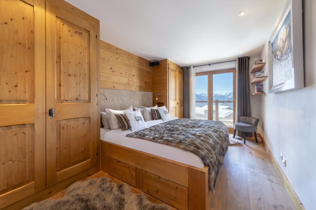 A cozy bedroom with wood-paneled walls and floors, a large bed with fur blankets, built-in wardrobes, a chair by a window with mountain views, and shelves above. Natural light fills the room. David Pearson Travel