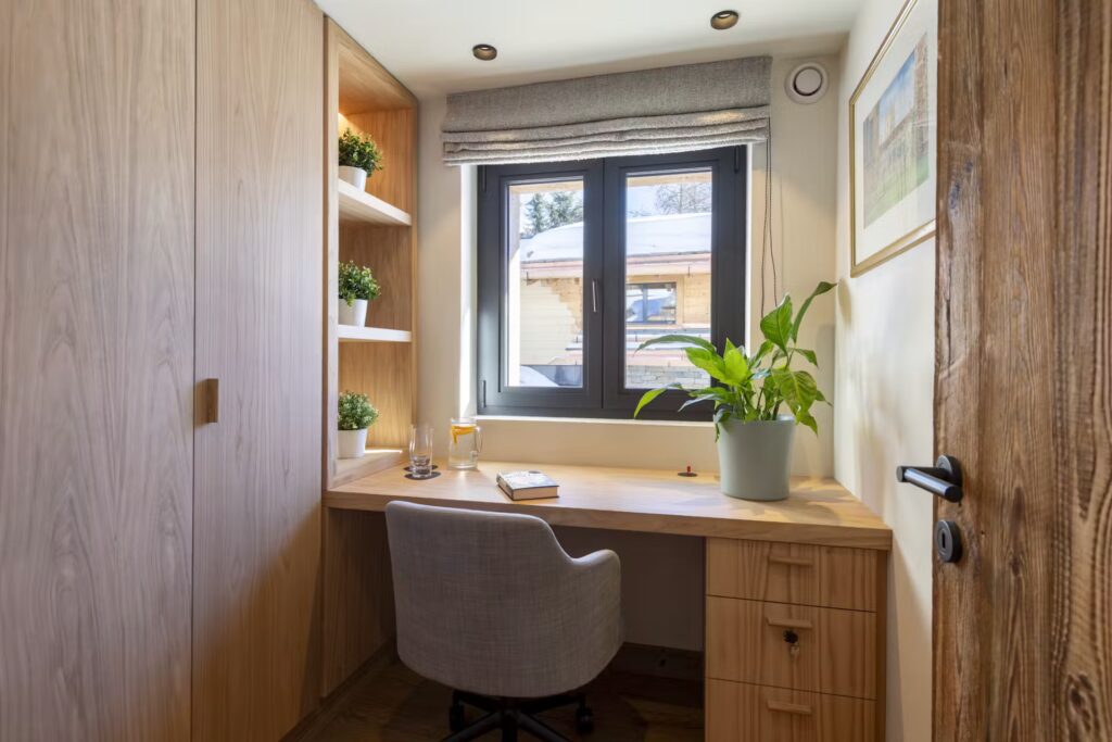 A small home office with a wooden desk, gray chair, potted plants, shelves with greenery, a notebook, and a window letting in natural light with a view of a wooden building and trees outside. David Pearson Travel