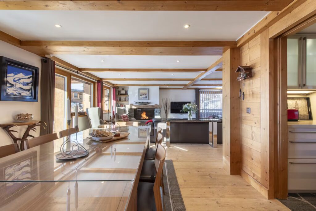 Open-plan living and dining area in a modern chalet with wooden beams, glass dining table, cozy seating by a fireplace, and large windows letting in natural light. Kitchen is visible to the right. David Pearson Travel